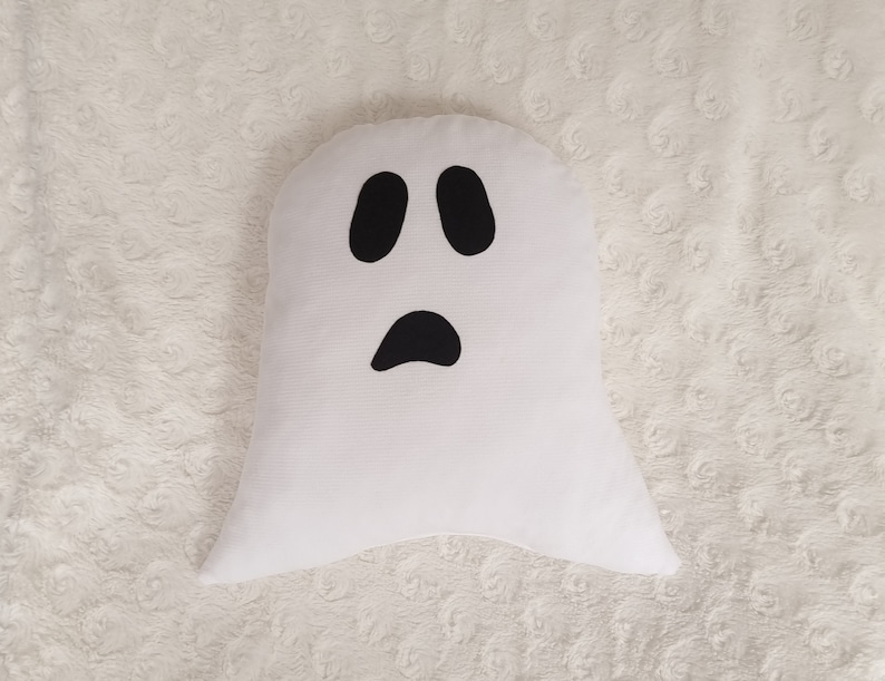 May include: White ghost-shaped pillow with a black face and two eyes. The pillow is on a white fuzzy surface.