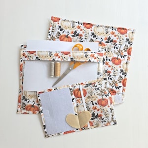 May include: A fabric organiser set featuring a pumpkin and foliage print in orange, brown, and black. The set includes a flat pouch with a clear window containing orange-handled scissors and two spools of thread, and a smaller folding case.