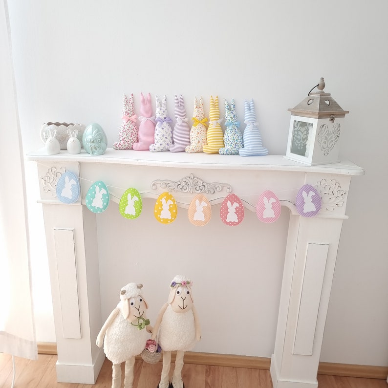 May include: A white mantelpiece with a pastel colored bunny garland and a set of pastel colored bunny figurines. The garland is made of felt and has a white bunny silhouette on each piece. The figurines are made of fabric and have a variety of pastel colors.
