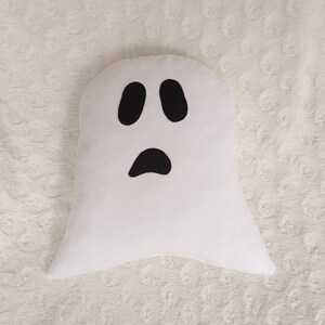 May include: White ghost-shaped pillow with a black face. The ghost has two black eyes and a black mouth.