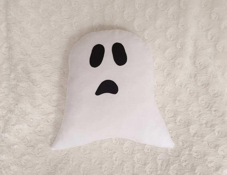 May include: White ghost-shaped pillow with a black face. The ghost has two black eyes and a black mouth.