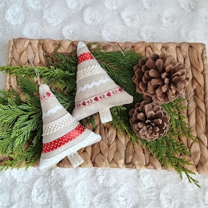 Könnte beinhalten: Zwei handgemachte Stoff-Weihnachtsbaumschmuckstücke mit rotem und weißem Besatz, auf einem geflochtenen Tablett mit Kiefernästen und Tannenzapfen.