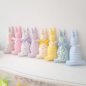 May include: A row of eight handmade plush bunny rabbits in various pastel colors with floral and striped patterns. The bunnies are arranged in a line on a white surface.
