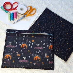 May include: A black Halloween-themed zippered pouch with a clear front panel, featuring a spooky print of skeletons, pumpkins, and a haunted house. A matching padded mat is next to the pouch. Crafting supplies, including scissors, ribbon, and thread, are also visible.