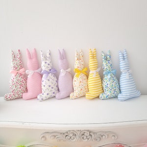 May include: Eight handmade fabric bunny rabbits in a rainbow of colors. The bunnies are lined up in a row on a white surface. They are all made of different patterned fabrics, including floral, striped, and polka dot.