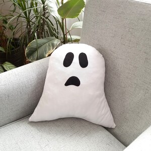May include: A white ghost-shaped pillow with a black face and two eyes. The pillow is sitting on a gray couch.