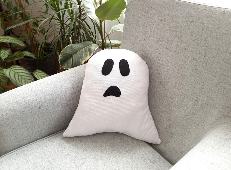 May include: A white ghost-shaped pillow with a black face and two eyes. The pillow is sitting on a gray couch.