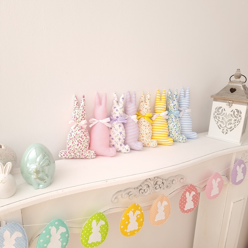 May include: A row of eight pastel-colored fabric bunnies with floral and striped patterns. The bunnies are arranged on a white shelf with a garland of pastel Easter eggs hanging below.