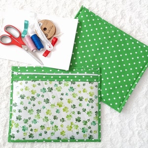 May include: Two green fabric pouches with white polka dots and a zipper closure. One pouch has a clear front with a white background and a green shamrock pattern. The pouches are on a white fuzzy surface with sewing supplies, including scissors, thread, and ribbon.