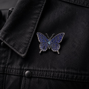 Puede incluir: Un broche de esmalte con forma de mariposa que presenta un fondo azul profundo con intrincados patrones celestiales en tono plateado, incluyendo estrellas y lunas crecientes. El centro de la mariposa muestra un peque&ntilde;o icono de luna creciente azul claro.