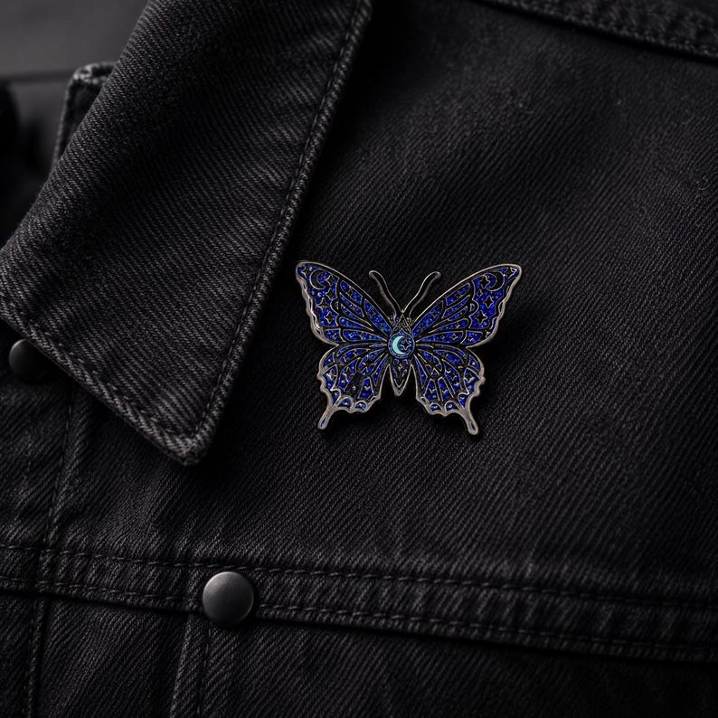 Puede incluir: Un broche de esmalte con forma de mariposa que presenta un fondo azul profundo con intrincados patrones celestiales en tono plateado, incluyendo estrellas y lunas crecientes. El centro de la mariposa muestra un peque&ntilde;o icono de luna creciente azul claro.