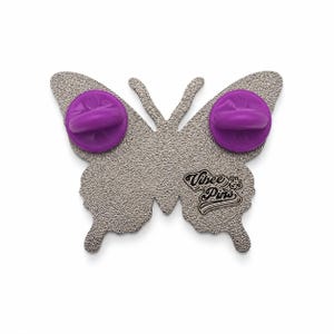 Puede incluir: Un broche de metal plateado con forma de mariposa y reverso texturizado. Dos cierres de goma morados est&aacute;n sujetos a los pines. El texto "Vibee Pins" est&aacute; grabado en la secci&oacute;n inferior del ala derecha del soporte met&aacute;lico con forma de mariposa.