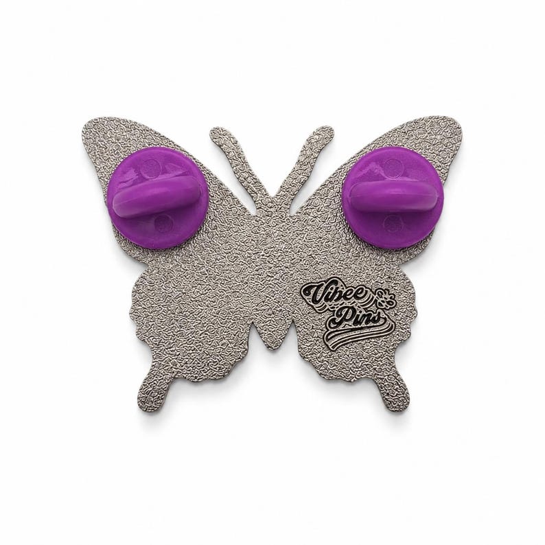 Puede incluir: Un broche de metal plateado con forma de mariposa y reverso texturizado. Dos cierres de goma morados est&aacute;n sujetos a los pines. El texto "Vibee Pins" est&aacute; grabado en la secci&oacute;n inferior del ala derecha del soporte met&aacute;lico con forma de mariposa.