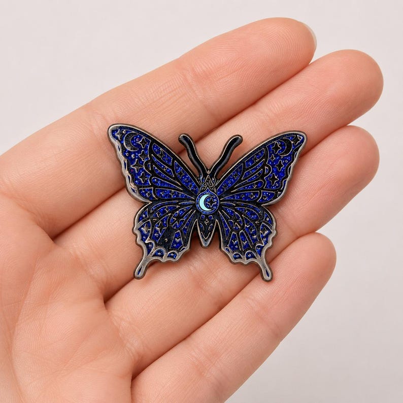 Puede incluir: Un broche de esmalte con forma de mariposa que presenta un acabado de purpurina azul oscuro con contornos negros. Las alas est&aacute;n decoradas con un patr&oacute;n celestial de estrellas y lunas crecientes, con una peque&ntilde;a luna azul claro en el centro.
