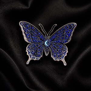 Puede incluir: Un broche de esmalte con forma de mariposa que presenta un acabado de purpurina azul oscuro con contornos de metal plateado. Las alas est&aacute;n decoradas con patrones celestiales que incluyen estrellas y lunas crecientes, con un emblema central.