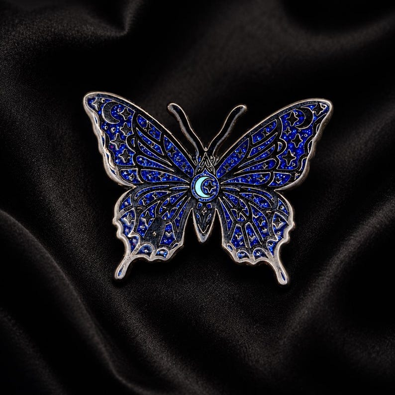 Puede incluir: Un broche de esmalte con forma de mariposa que presenta un acabado de purpurina azul oscuro con contornos de metal plateado. Las alas est&aacute;n decoradas con patrones celestiales que incluyen estrellas y lunas crecientes, con un emblema central.