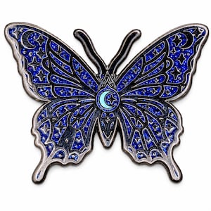 Puede incluir: Un broche de esmalte con forma de mariposa que presenta un patr&oacute;n de cielo nocturno con purpurina azul oscuro y negro, con contornos de metal plateado. Las alas est&aacute;n decoradas con motivos celestiales de estrellas y lunas crecientes, con una luna central.