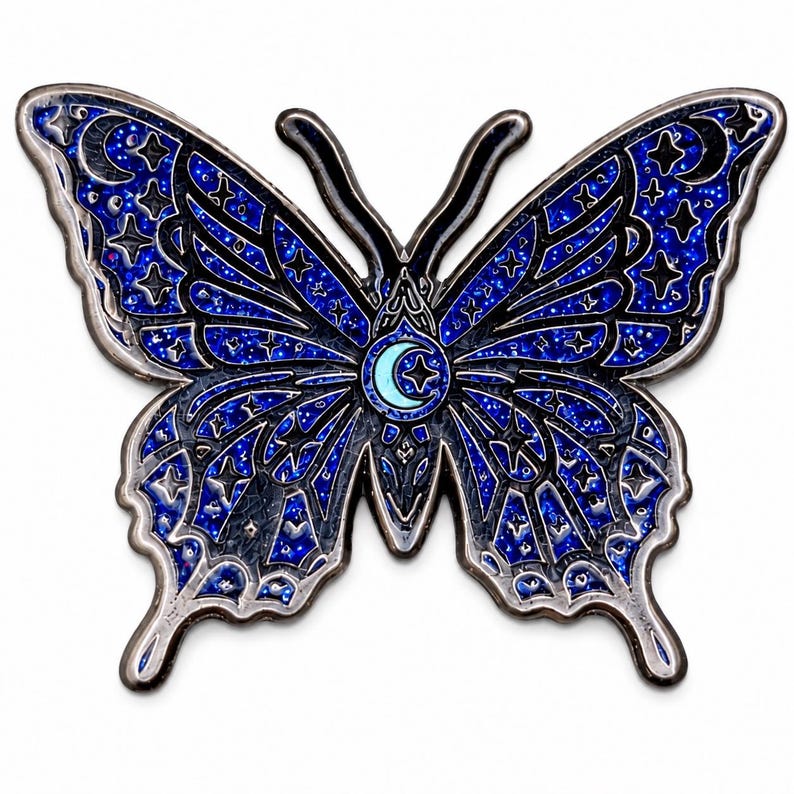 Puede incluir: Un broche de esmalte con forma de mariposa que presenta un patr&oacute;n de cielo nocturno con purpurina azul oscuro y negro, con contornos de metal plateado. Las alas est&aacute;n decoradas con motivos celestiales de estrellas y lunas crecientes, con una luna central.