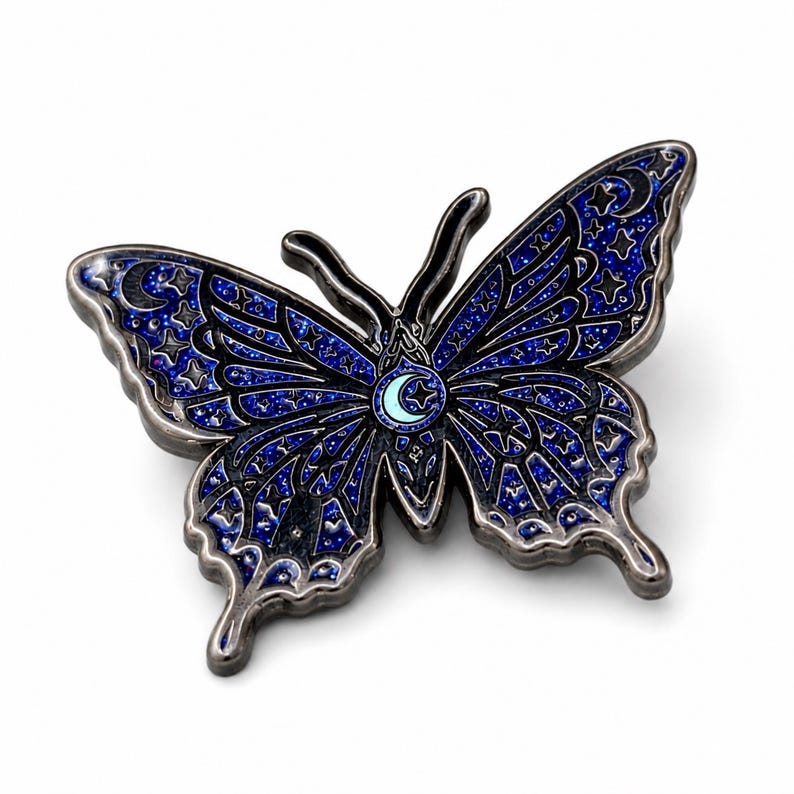 Puede incluir: Un broche de esmalte con forma de mariposa que presenta un fondo azul oscuro brillante con contornos negros. Las alas est&aacute;n decoradas con patrones celestiales que incluyen estrellas y lunas crecientes. Una peque&ntilde;a luna creciente azul claro est&aacute; en el centro.