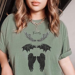 May include: Sage green t-shirt featuring three sets of stylized wings in black ink. The top set of wings is feathered, the middle set is bat-like, and the bottom set is solid black. The text "Time to Fly" is above the top wings.