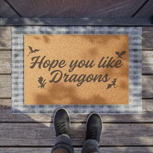 May include: A brown coir doormat with a white checkered border. The doormat has the text "Hope you like Dragons" printed on it in black. There are also several black dragon silhouettes on the doormat.