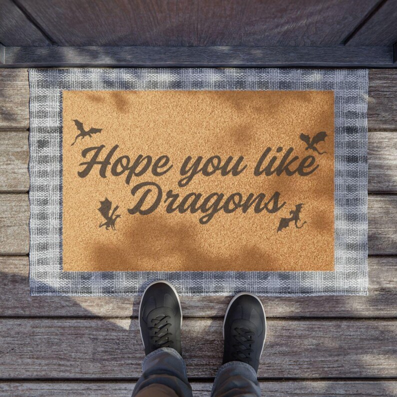 May include: A brown coir doormat with a white checkered border. The doormat has the text "Hope you like Dragons" printed on it in black. There are also several black dragon silhouettes on the doormat.