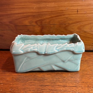 May include: A rectangular, light blue ceramic planter with a leaf design. The planter has a textured surface and a scalloped rim. The interior is smooth and the exterior features a repeating leaf pattern.