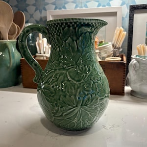May include: A green ceramic pitcher featuring a raised relief pattern of overlapping maple leaves across its body. The vessel has a curved handle, a wide spout, and a checkered texture along the upper rim, finished with a glossy glaze.