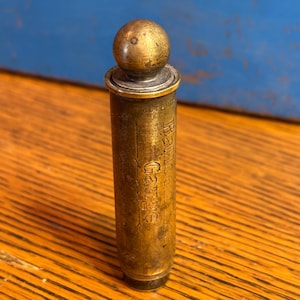 May include: A vintage brass container with a rounded top and a small sphere finial. The cylindrical body features the engraved text "Ball Garage". The brass has a warm, aged patina, suggesting its antique nature. The container is standing upright.