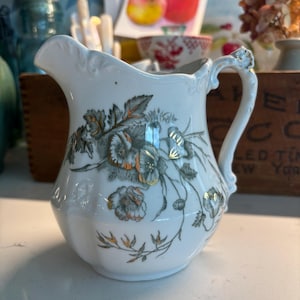 May include: A white porcelain creamer pitcher featuring a decorative floral pattern in shades of grey and green with gold metallic accents. The pitcher has a scalloped rim, a curved handle, and a rounded body with embossed detailing near the top.