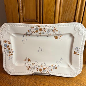 May include: A rectangular white serving platter with a floral design. The platter has a scalloped edge with embossed shell-like details at the corners. The floral pattern includes brown leaves and small blue flowers. The platter is supported by a silver metal stand.