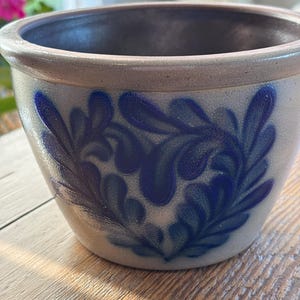 May include: A ceramic bowl with a light beige exterior and a dark blue interior. The bowl features a blue floral design on the exterior. The rim is a lighter beige color. The bowl is sitting on a wooden surface.
