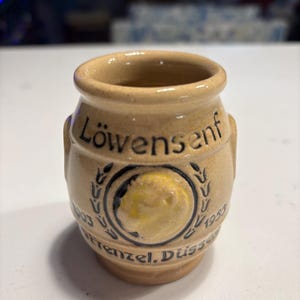 May include: A small, tan ceramic jar with the word "Löwensenf" printed on the front. The jar has a decorative emblem with the years 1903 and 1953. The jar has a small handle on each side.