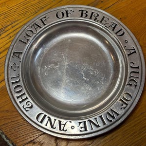 May include: A round, silver-colored metal plate with the embossed text "Thou A Loaf Of Bread A Jug Of Wine And". The plate has a shallow, concave center and a wide, raised rim with the text. The plate is sitting on a wooden surface.