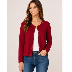 May include: A burgundy cardigan with a round neckline and long sleeves. The cardigan has a sequined trim along the neckline, button placket, and cuffs. The buttons are small and silver. The person is wearing blue jeans and a white top.