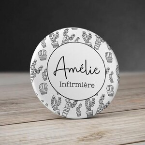 Puede incluir: Un botón redondo blanco con un diseño de cactus en blanco y negro. El círculo central presenta el nombre "Amélie" en cursiva y la palabra "Infirmière" debajo. El botón está sobre una superficie de madera.