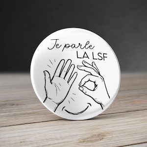 May include: A circular white pinback button featuring a black line drawing of two hands and a smiling mouth. The text "Je parle LA LSF" is printed in black above the hands. The design includes a hand with fingers spread and a hand forming a circle.