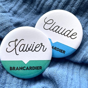 Può includere: Due bottoni rotondi bianchi con testo nero. Il bottone superiore dice "Claude" con testo blu sotto che dice "Brancardier". Il bottone inferiore dice "Xavier" con testo verde sotto che dice "Brancardier".