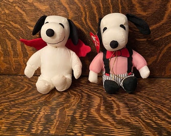 Può includere: Due peluche di Snoopy. Uno è vestito da vampiro con un mantello rosso e l'altro è vestito da gentiluomo con una camicia rosa, bretelle nere e un papillon.