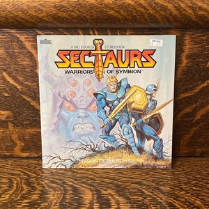 May include: A vintage paperback storybook titled "Sectaurs: Warriors of Symbion" with a cover illustration of three characters in a fantasy setting. The cover features a blue and gold warrior, a blue warrior with a sword, and a large, menacing creature with glowing red eyes.