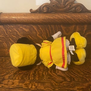 May include: A yellow plush toy with a red and white striped sash and a yellow hat. The toy is lying on a wooden surface.