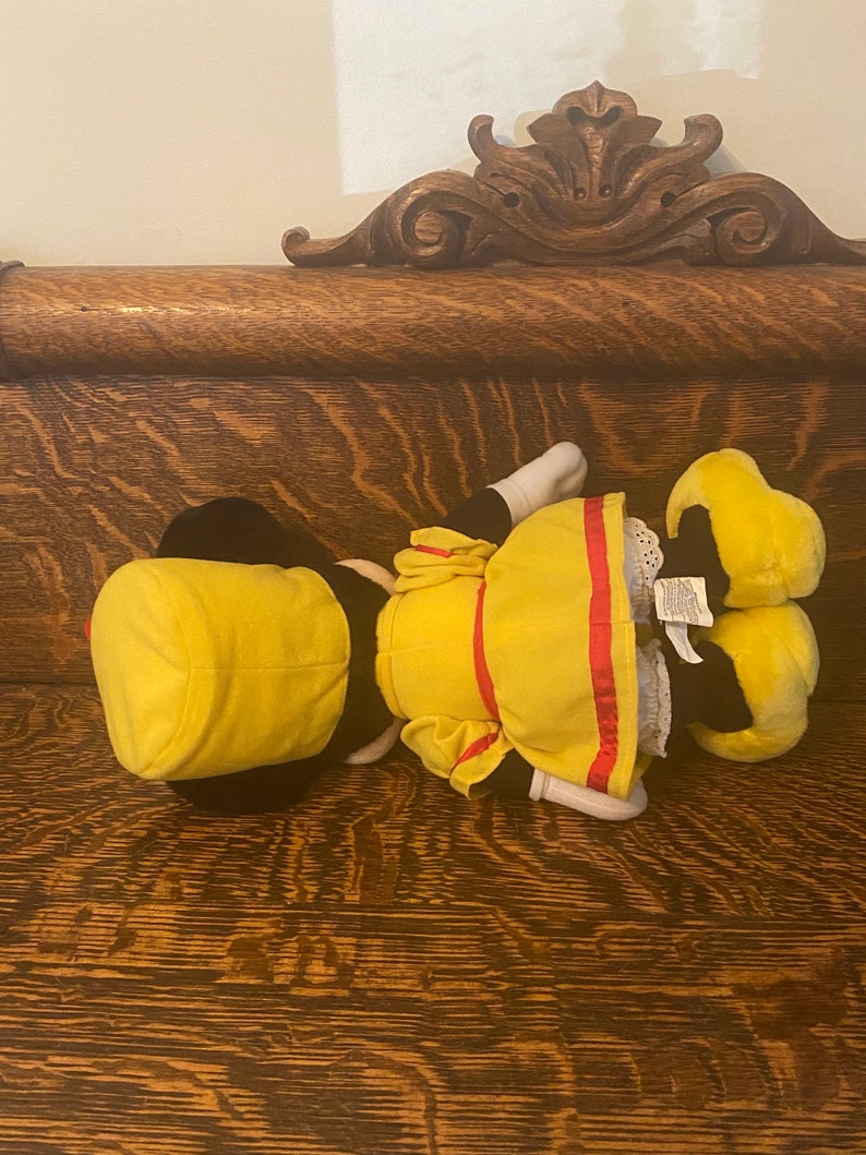 May include: A yellow plush toy with a red and white striped sash and a yellow hat. The toy is lying on a wooden surface.
