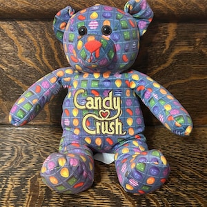 Puede incluir: Un oso de peluche con un fondo azul y un colorido patrón de caramelos. El oso tiene ojos negros, una nariz roja y las palabras "Candy Crush" bordadas en el pecho. El oso está sentado sobre una superficie de madera.