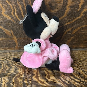 Op de afbeelding: Een knuffel van een tekenfilmmuis met zwarte oren, een roze strik en een roze jurk. Het personage heeft een cr&egrave;mekleurig gezicht, zwarte ledematen en witte handschoenen. Een doorzichtig plastic labelbevestigingsmiddel zit aan het hoofd.