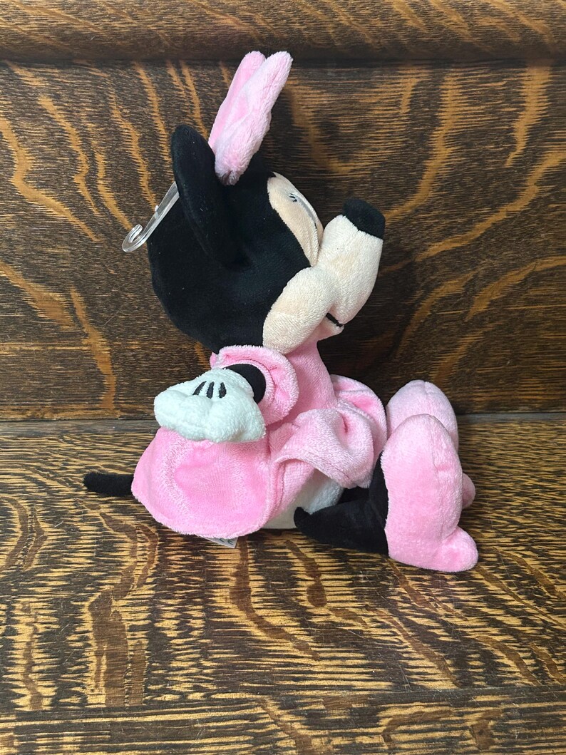 Op de afbeelding: Een knuffel van een tekenfilmmuis met zwarte oren, een roze strik en een roze jurk. Het personage heeft een cr&egrave;mekleurig gezicht, zwarte ledematen en witte handschoenen. Een doorzichtig plastic labelbevestigingsmiddel zit aan het hoofd.