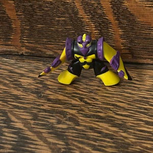 May include: A yellow, black, and purple action figure in a crouching pose. The figure has a robot-like design with articulated limbs and a detailed head. The figure is set against a wooden background.