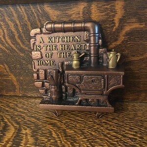 May include: A brown metal wall hanging with a stove design and the text "A kitchen is the heart of the home".