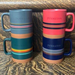 Puede incluir: Cuatro tazas de cerámica apiladas en dos columnas. La superior izquierda es gris, la inferior izquierda es verde azulado, la superior derecha es coral y la inferior derecha es azul marino. Cada taza tiene la palabra PENDLETON, rayas y base de terracota.