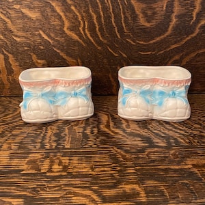 May include: Two white ceramic planters shaped like baby shoes with blue bows and pink trim.