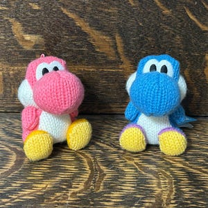 May include: Two small plush figures, one pink and one blue, each featuring a large rounded snout, white belly, and yellow feet. These knitted-style characters have black and white eyes and are positioned side-by-side in a seated posture.